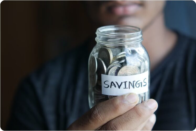 손에 들린 투명 유리병에 SAVINGS 라벨이 붙어 동전이 가득 담긴 모습